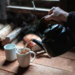 Rustic scene of hot tea being poured from a teapot into a mug by the window.