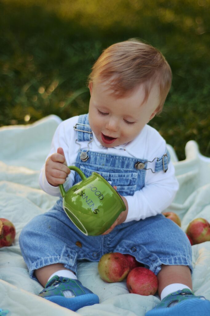 Cute toddler in denim jumper sitting on blanket outdoors holding a green mug.
